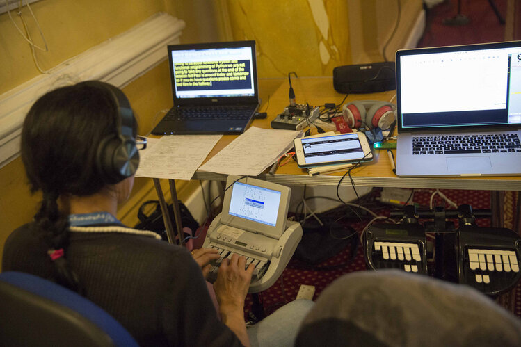 A person with headphones and a special stenographic keyboard, sitting in front of a desk with several papers and laptops.